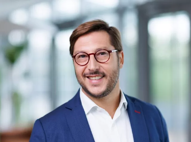 Headshot of Alexandre Cotarmanac’h, a man with brown hair, wearing red circular glasses, a blue blazer and a white dress shirt smiling with teeth